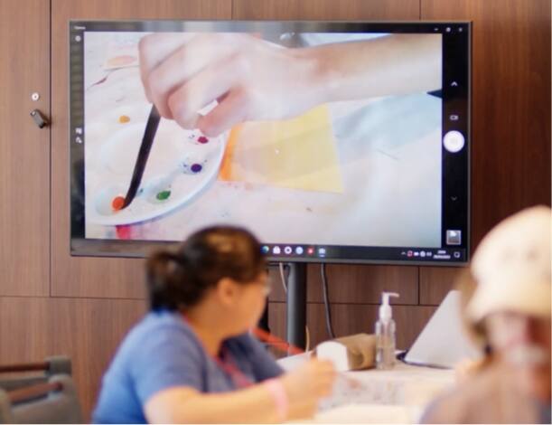 A woman in an art class looking at a monitor showing someone painting.