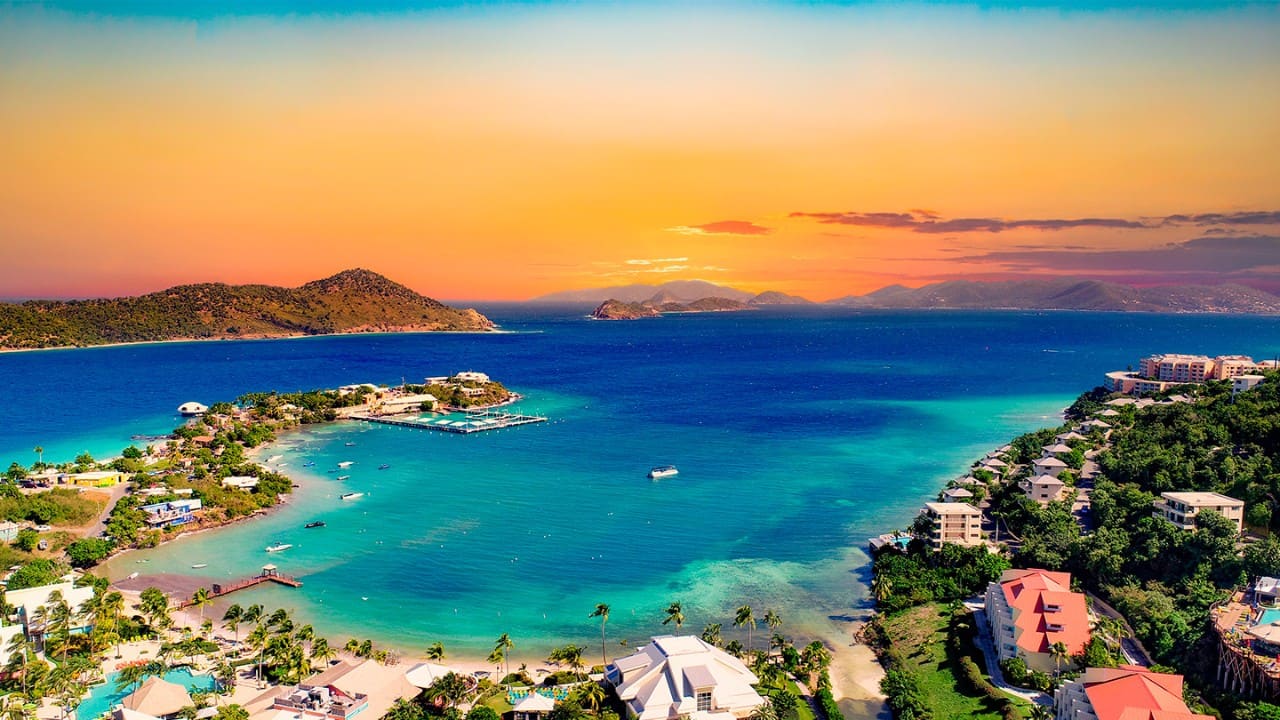 Aerial view of St. Thomas, U.S. Virgin Islands, featuring turquoise Caribbean waters, a tropical bay, waterfront resorts, palm-lined coastline, and a colorful sunset over the island landscape.