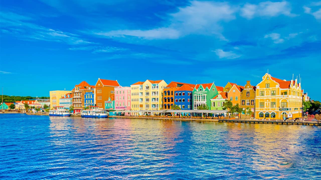 Colorful waterfront buildings lining the harbor in Willemstad, Curaçao, reflected in calm blue water beneath a dramatic sky with scattered clouds.