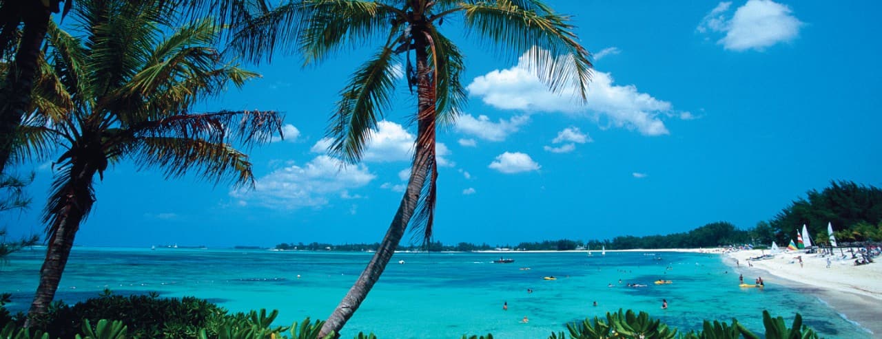 Palm trees framing a white-sand beach and turquoise waters in Nassau, Bahamas, with people swimming and relaxing along the shoreline.