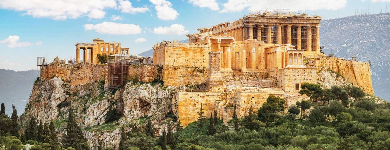 Panoramic view of the Acropolis of Athens with the Parthenon and surrounding ancient stone ruins atop a rocky hill, under a blue sky with scattered clouds.