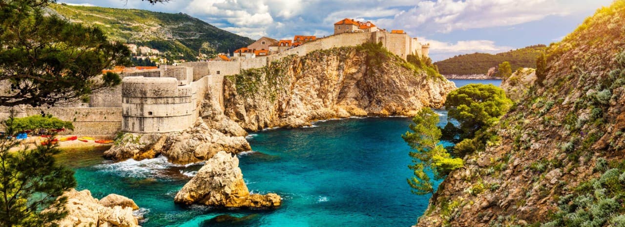 Panoramic view of Dubrovnik Old Town Croatia with stone fortress walls on rugged cliffs above a turquoise Adriatic Sea cove, Mediterranean coastline landscape.