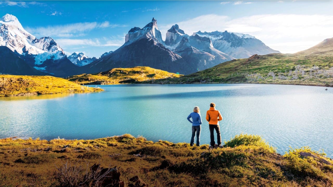 Couple stand beside glacial lake looking towards ice-capped mountains at Torres del Paine National Park in Patagonia, Chile.