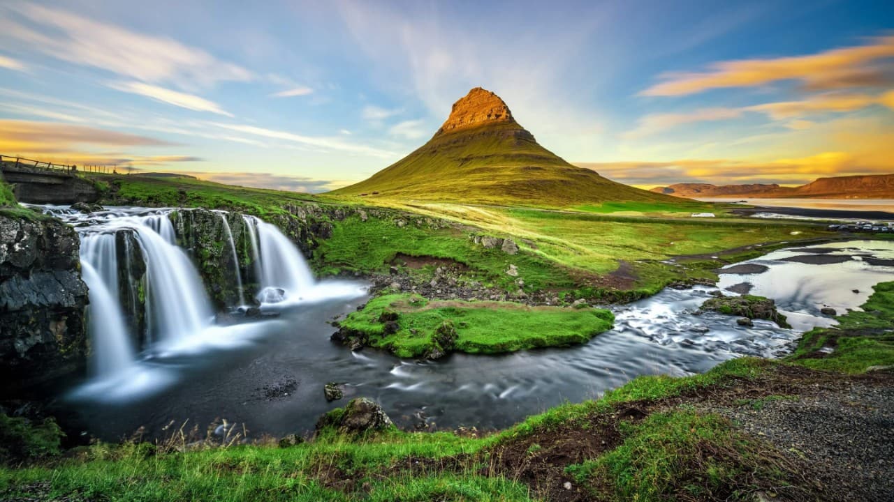 Pointed mountain, waterfall, and river in a lush Icelandic landscape at sunrise or sunset.