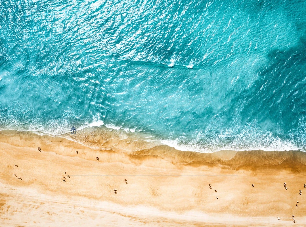 Aerial view looking down at a sunny Mediterranean beach with azure blue waters.
