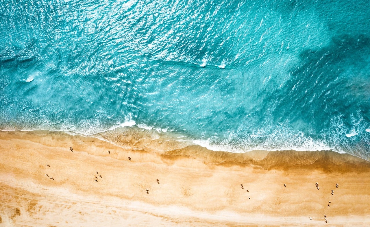 Aerial view looking down at a sunny Mediterranean beach with azure blue waters.