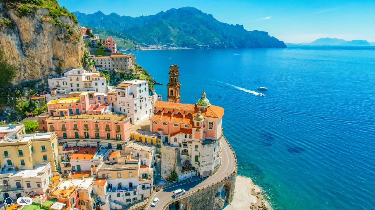 Cliffside coastal town with pastel buildings, a domed church, winding coastal road, and deep blue sea stretching toward distant mountains.