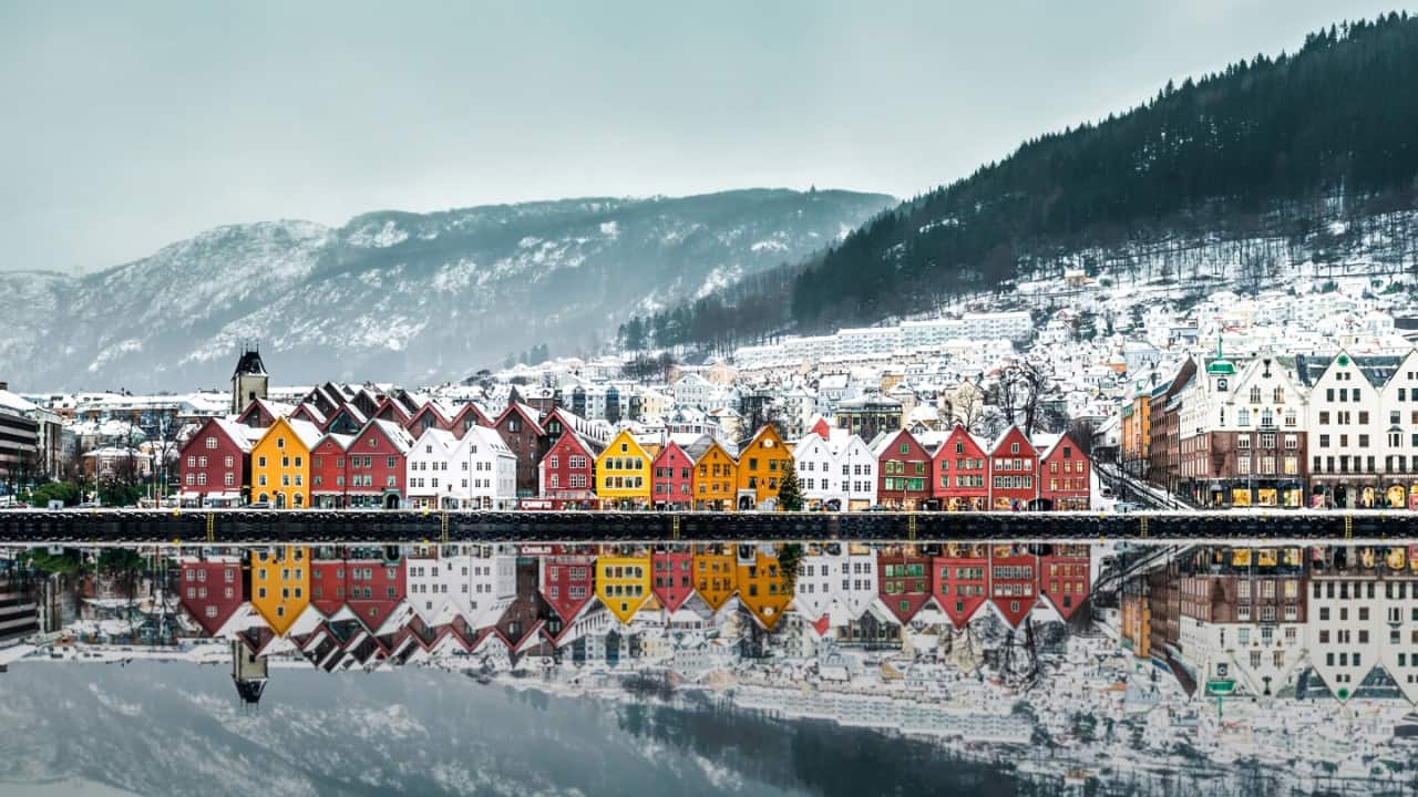 Colorful wooden houses lined along a waterfront, reflected in calm water, with snow-covered hills and mountains in the background.