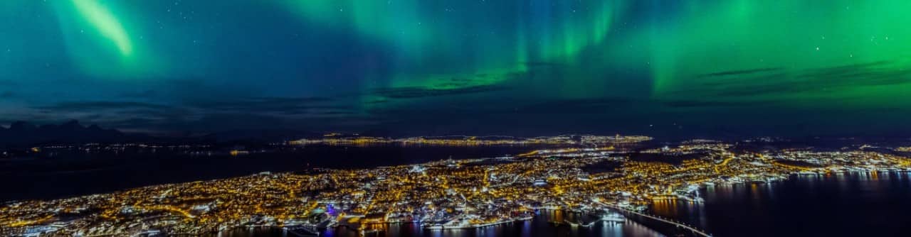 Aerial view of a coastal city at night illuminated by streetlights, with green northern lights glowing across the dark sky above the water