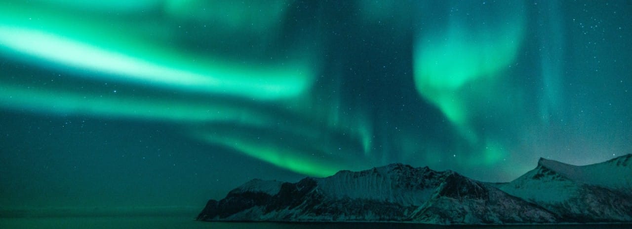 Aurora borealis illuminating the night sky above snow-covered mountains and a coastal landscape in Norway, showcasing vibrant green northern lights, Arctic scenery, and winter travel destination.