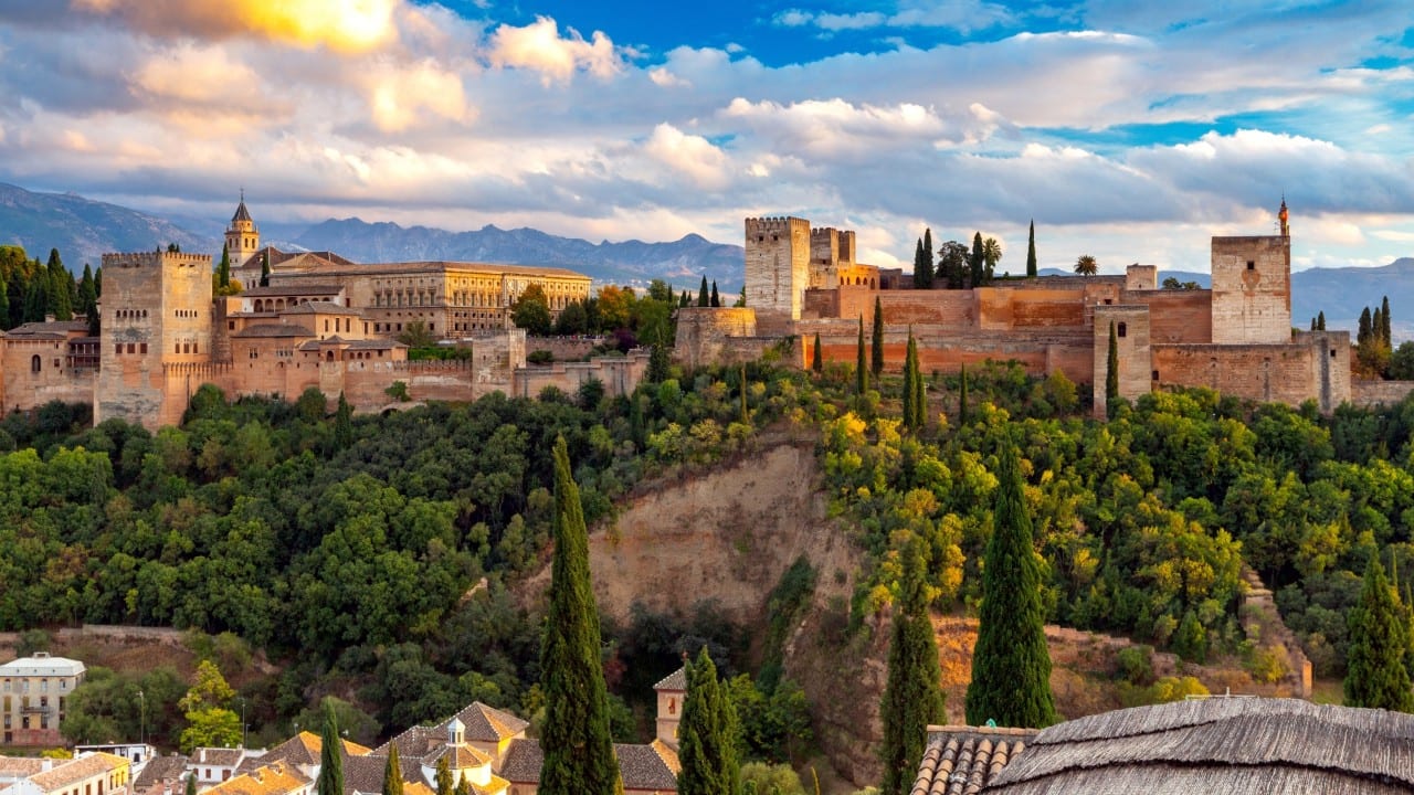 Alhambra Palace complex in Granada overlooking wooded hills, with historic towers, walls, and mountain views in the background.
