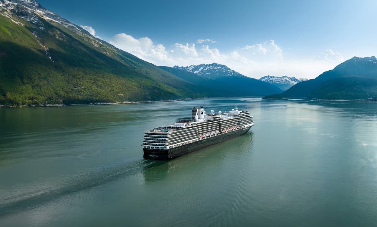 The Nieuw Amsterdam cruise ship sailing through a calm fjord surrounded by snow-capped mountains.