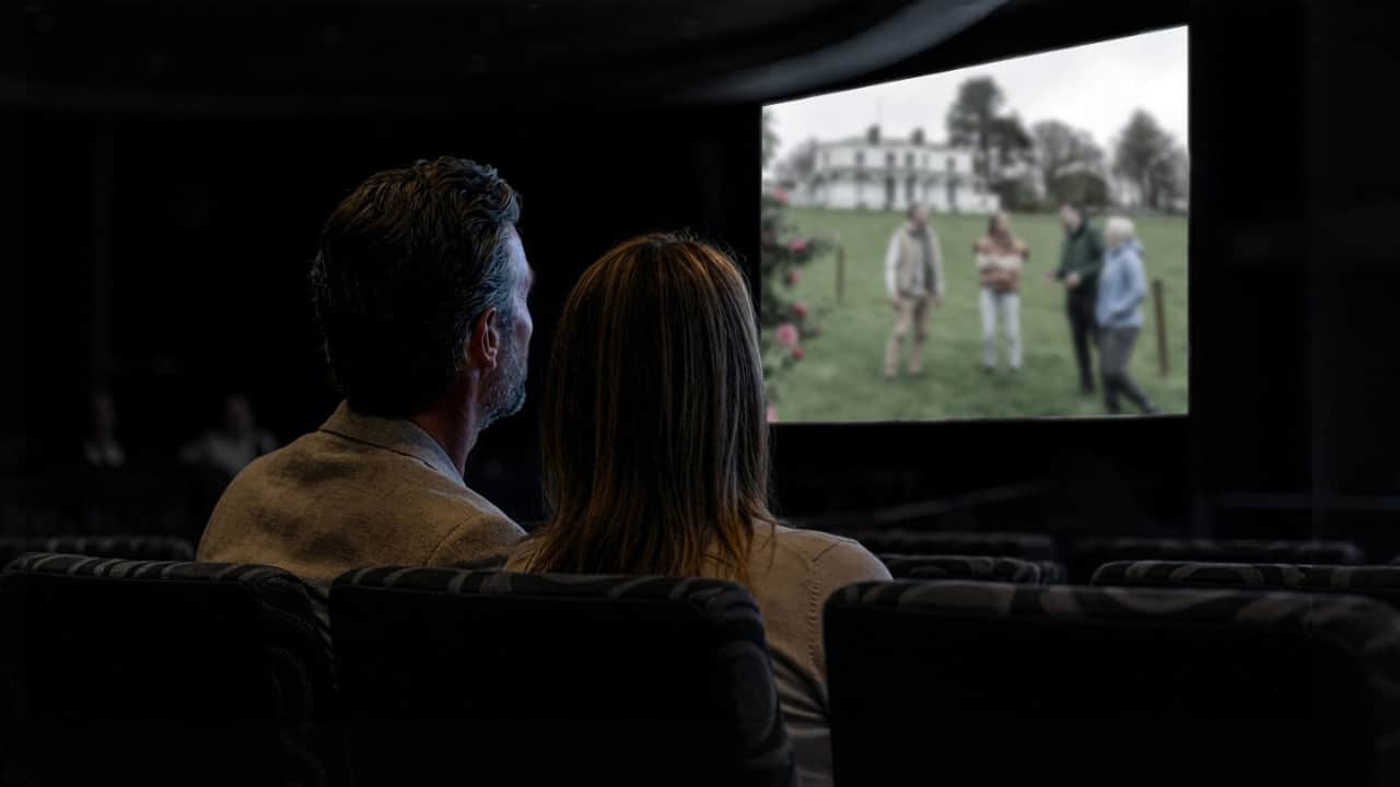 Two people sitting in a dark movie theater watching a film on a large screen showing a blurred outdoor scene with several people near a house.