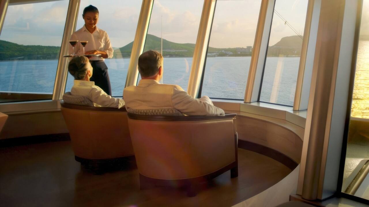 Guests seated by panoramic windows in an observation lounge aboard a cruise ship while a server delivers drinks overlooking the ocean and coastline.