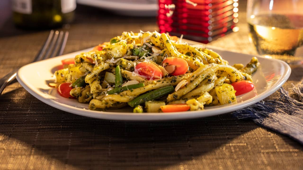 Plate of pesto pasta with green beans, cherry tomatoes, grated cheese, and sliced almonds served on a dining table, with a glass of white wine in the background.