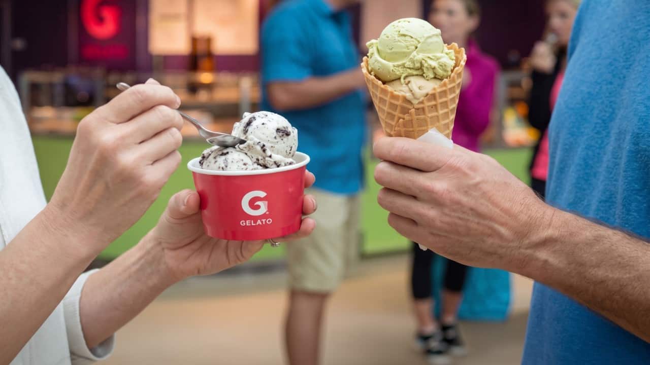 Two servings of gelato, one in a branded cup and one in a waffle cone, held by guests in an outdoor cruise ship setting.