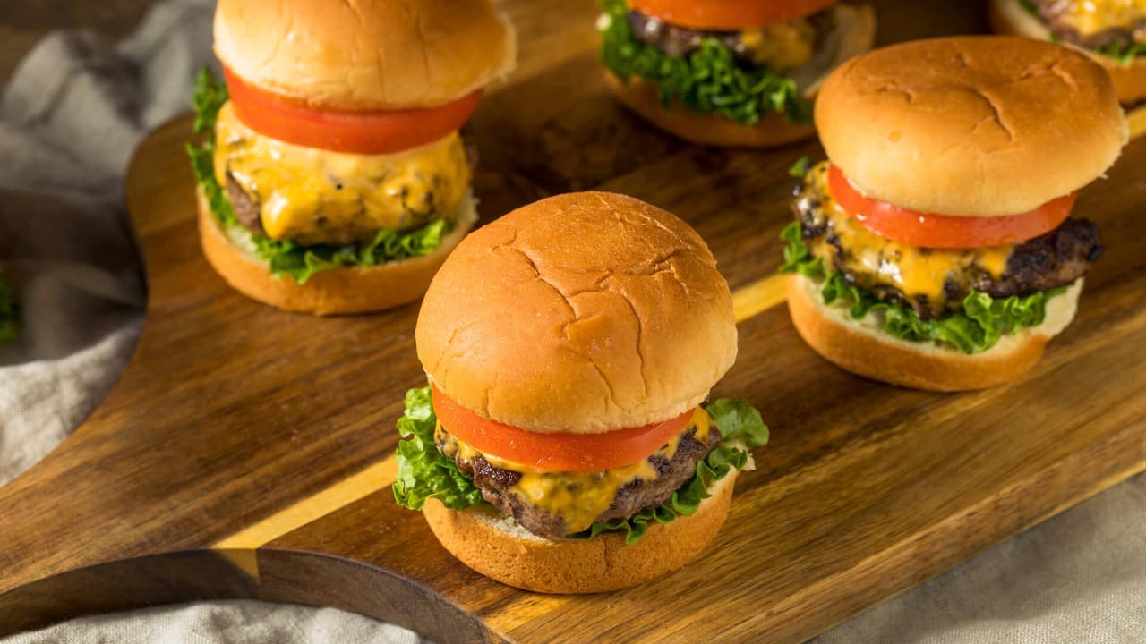 Four cheeseburger sliders topped with lettuce, melted cheese, and tomato slices, arranged on a wooden serving board.