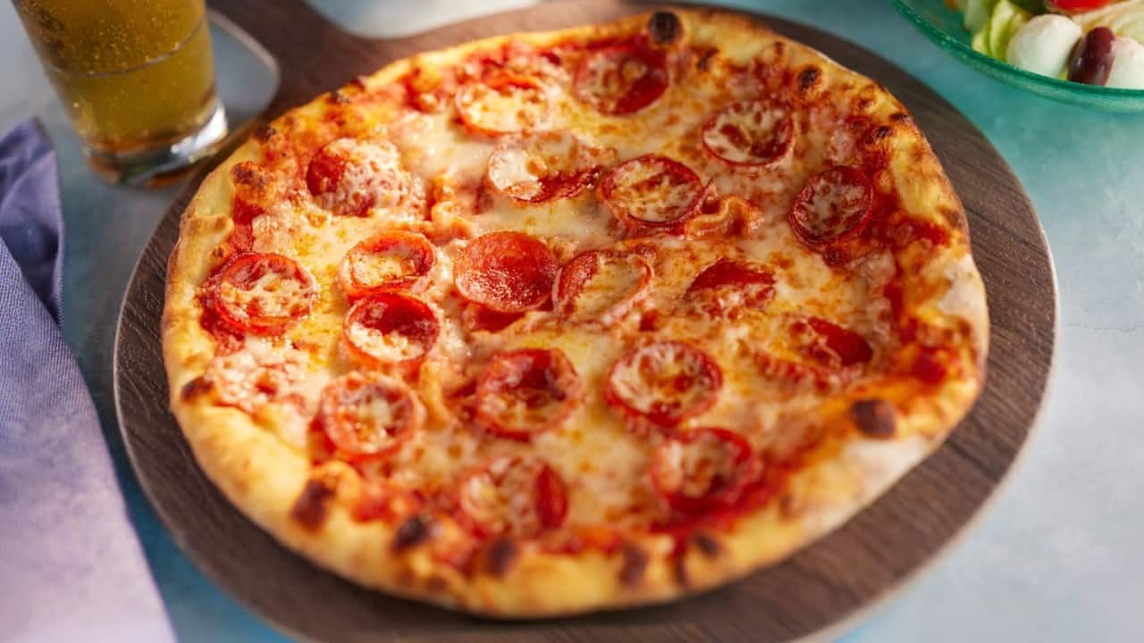 Pepperoni pizza with melted cheese served on a wooden board beside a drink.