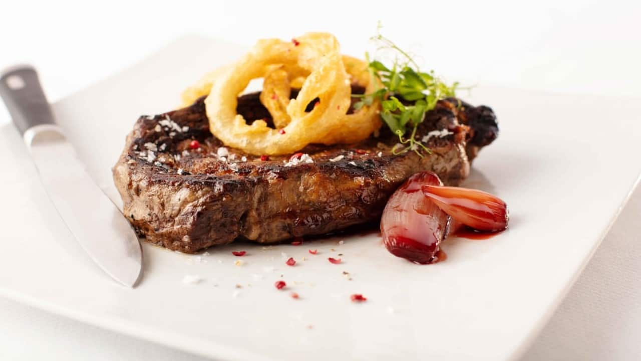 Grilled ribeye steak topped with crispy onion rings, sea salt, and peppercorns, served with glazed shallots on a minimalist white plate, gourmet steakhouse fine dining dish.