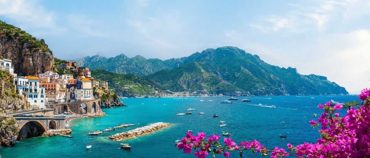 Coastal village perched on cliffs above a turquoise bay, with boats on the water, green mountains in the background, and vibrant pink flowers in the foreground.