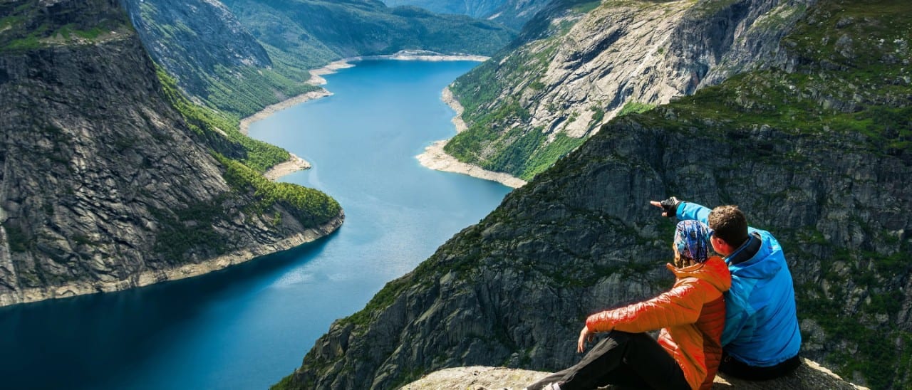 Two hikers sit on a rocky cliff overlooking a winding blue fjord surrounded by steep mountain slopes.