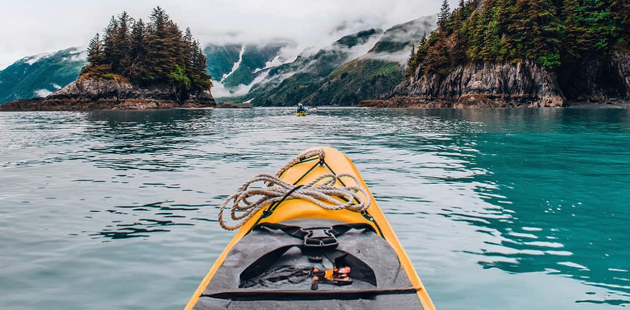 A kayak on turquoise water in Kenai Fjords National Park, Alaska, surrounded by forested mountains under a cloudy sky.
