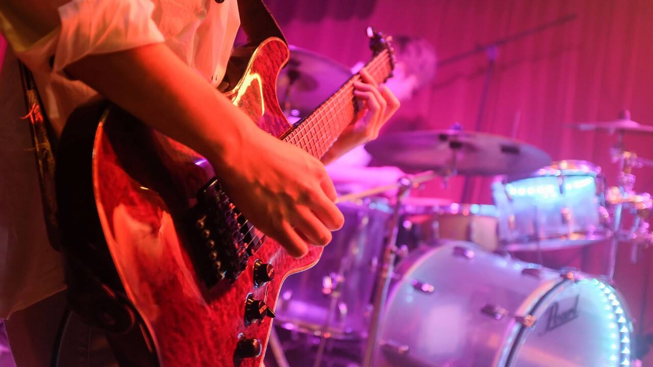 Close-up of a guitarist's hand playing a red electric guitar on stage, drum kit visible in the background.